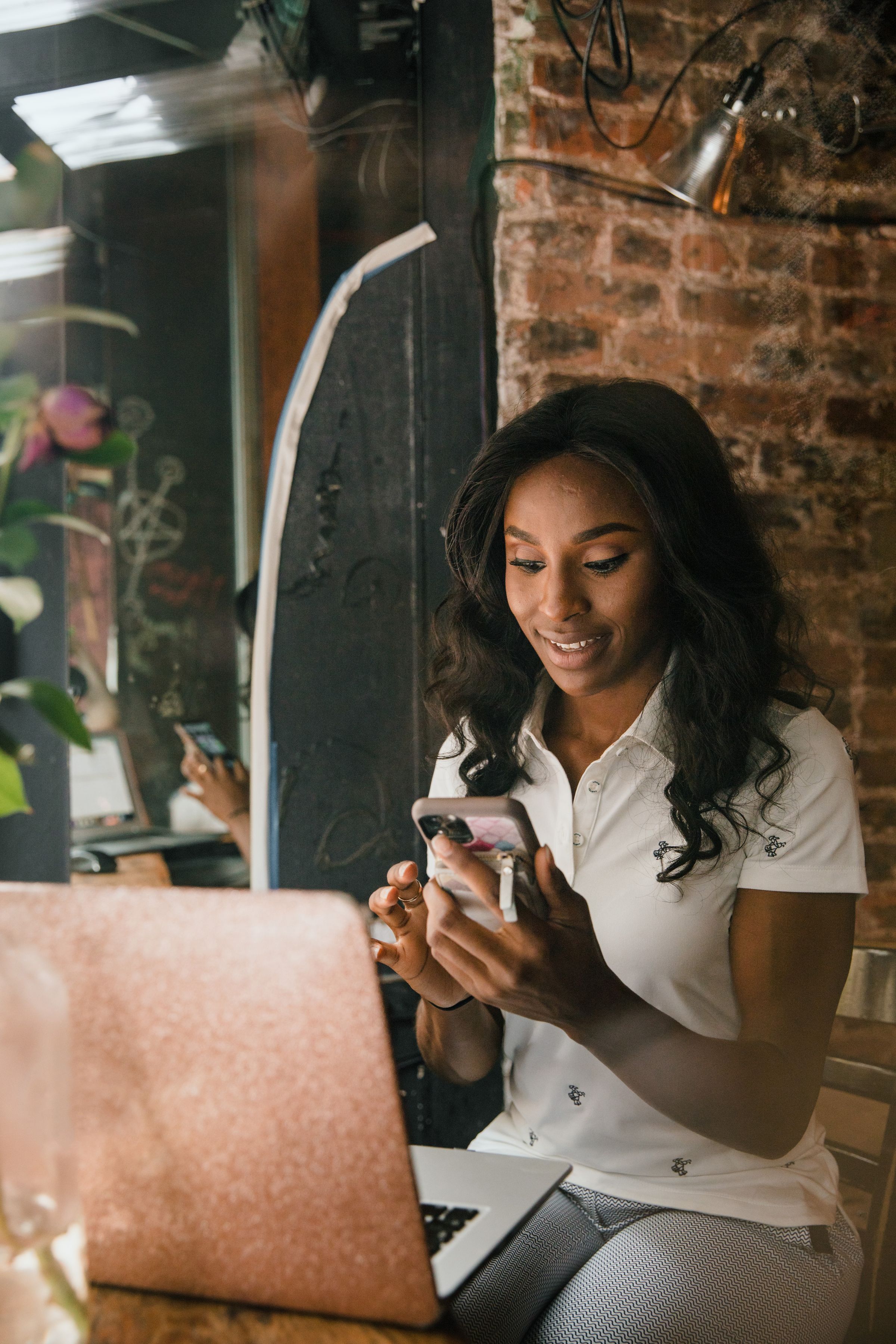 A woman smiling at her phone beside her laptop in a café