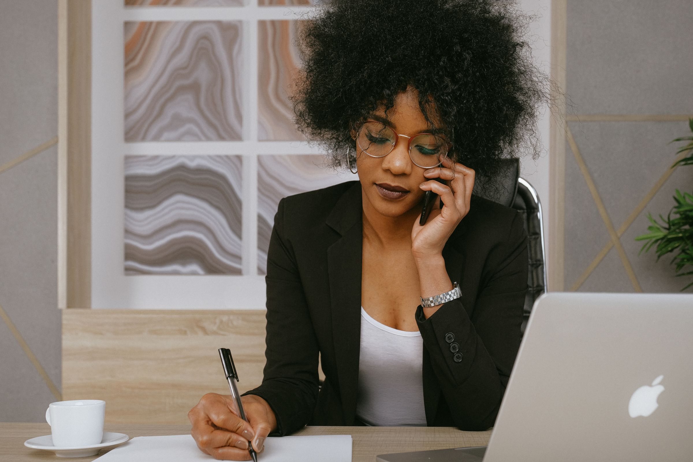 A woman in a blazer on a phone call, taking notes at her desk beside a laptop