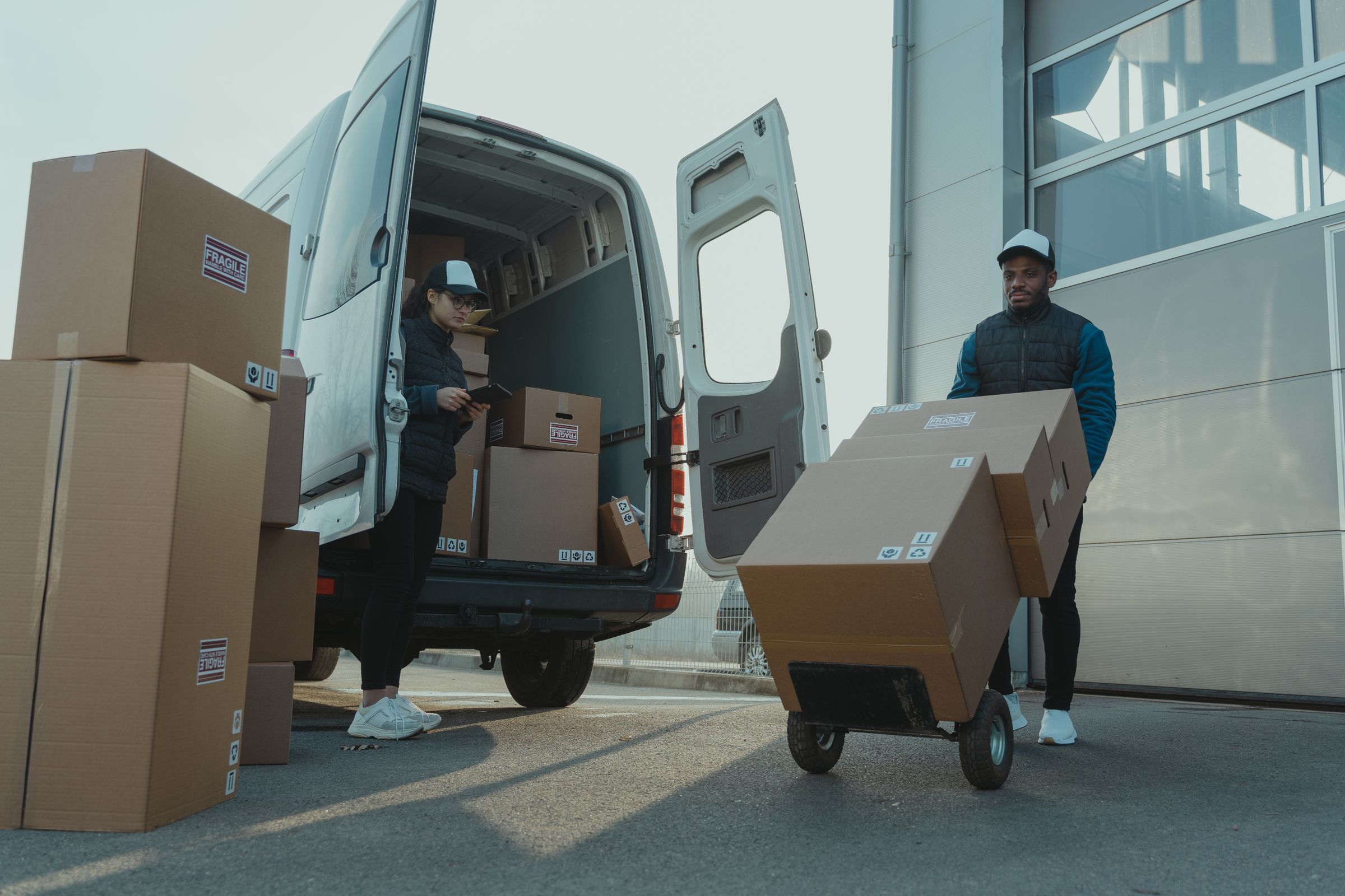 A courier wheeling boxes to a delivery van while a colleague checks a tablet