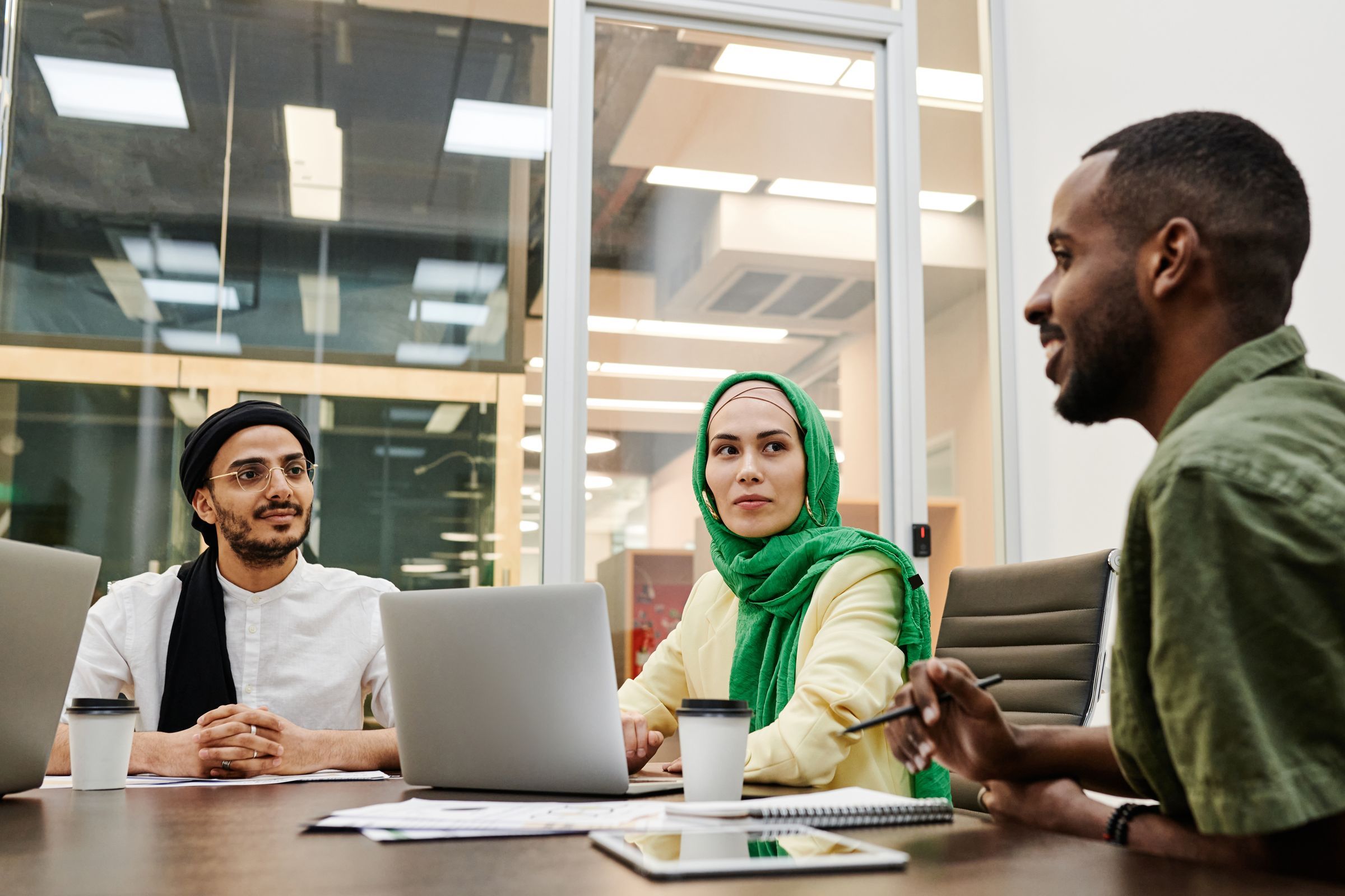 Three colleagues talking around a table with laptops in a glass-walled office